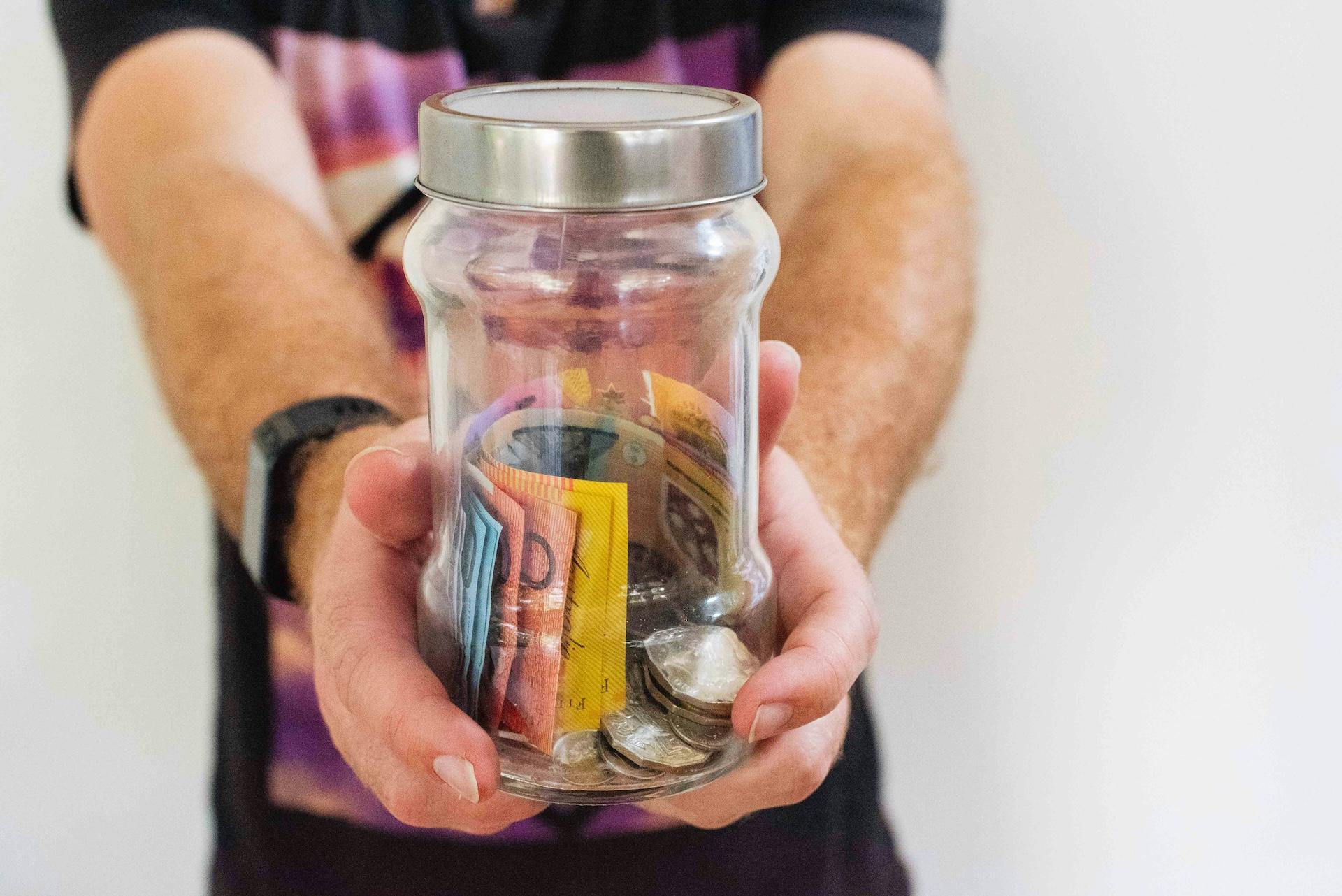 Glass money jar with colorful banknotes and coins being held in hands, representing personal savings and peer-to-peer investment opportunities