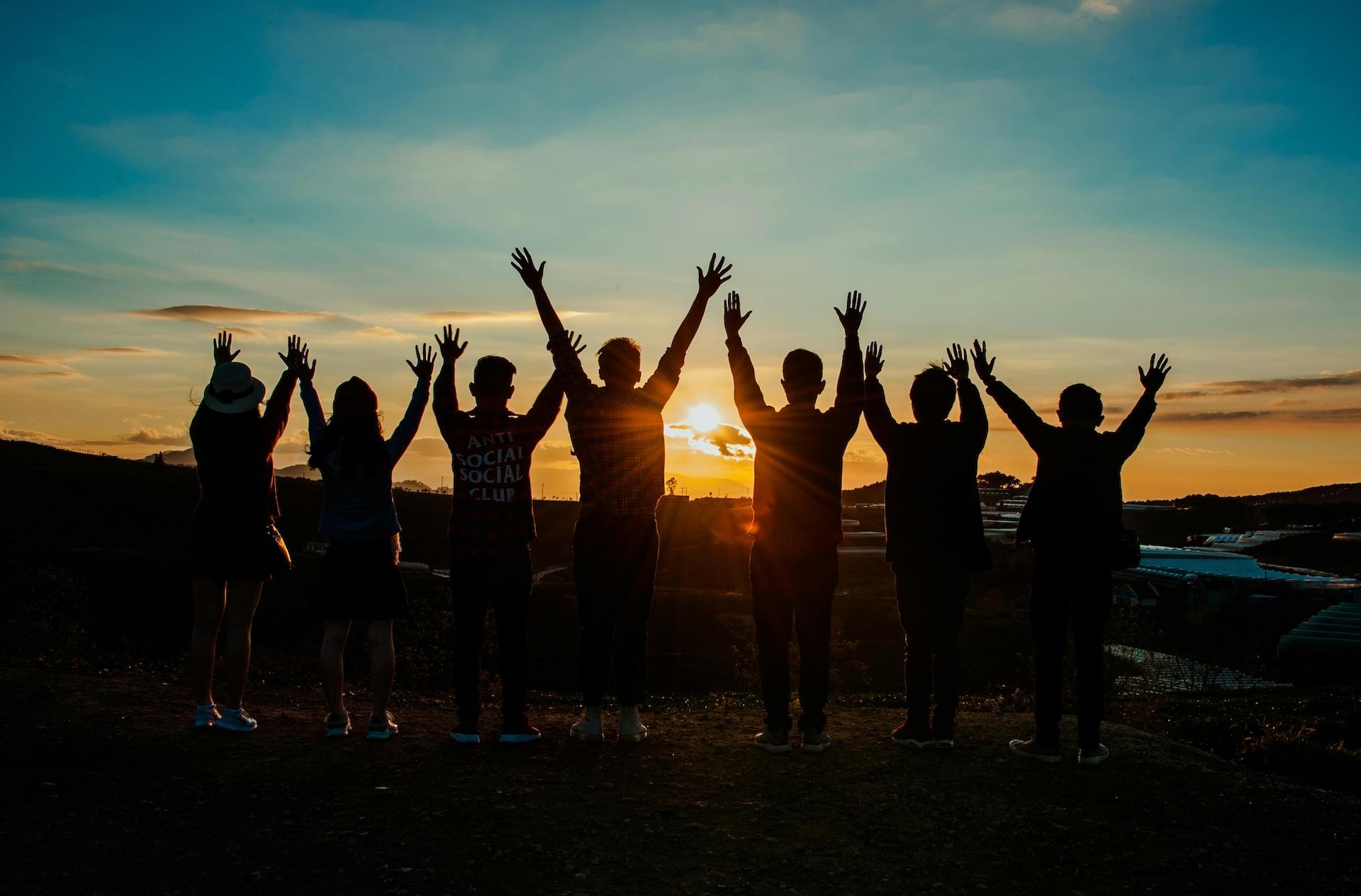 Silhouettes of people raising hands together against sunset sky