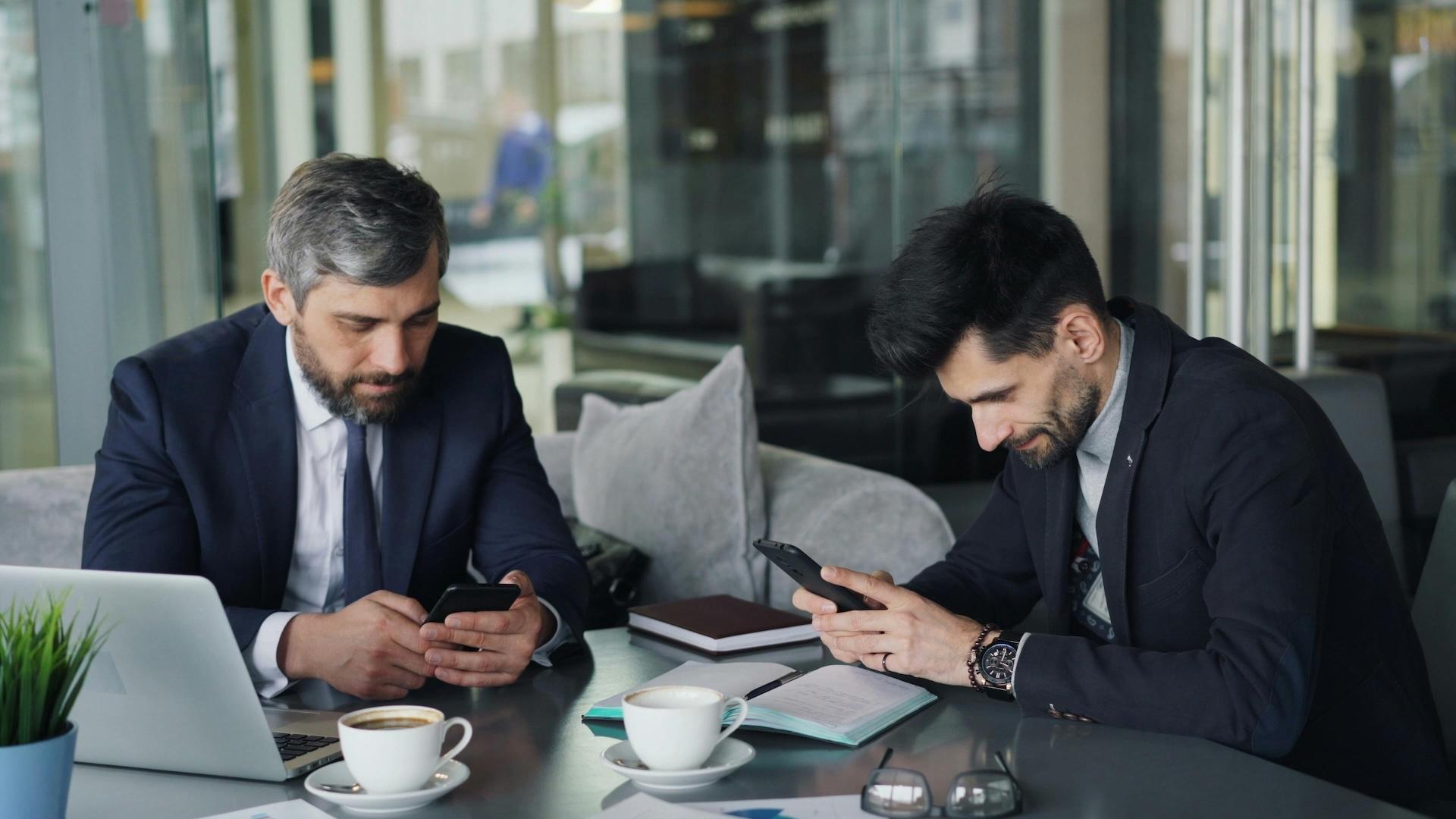 Two businessmen in suits sitting at a modern office table, focused on their smartphones. The table features notebooks, a laptop, and coffee cups, with a professional environment visible in the background.