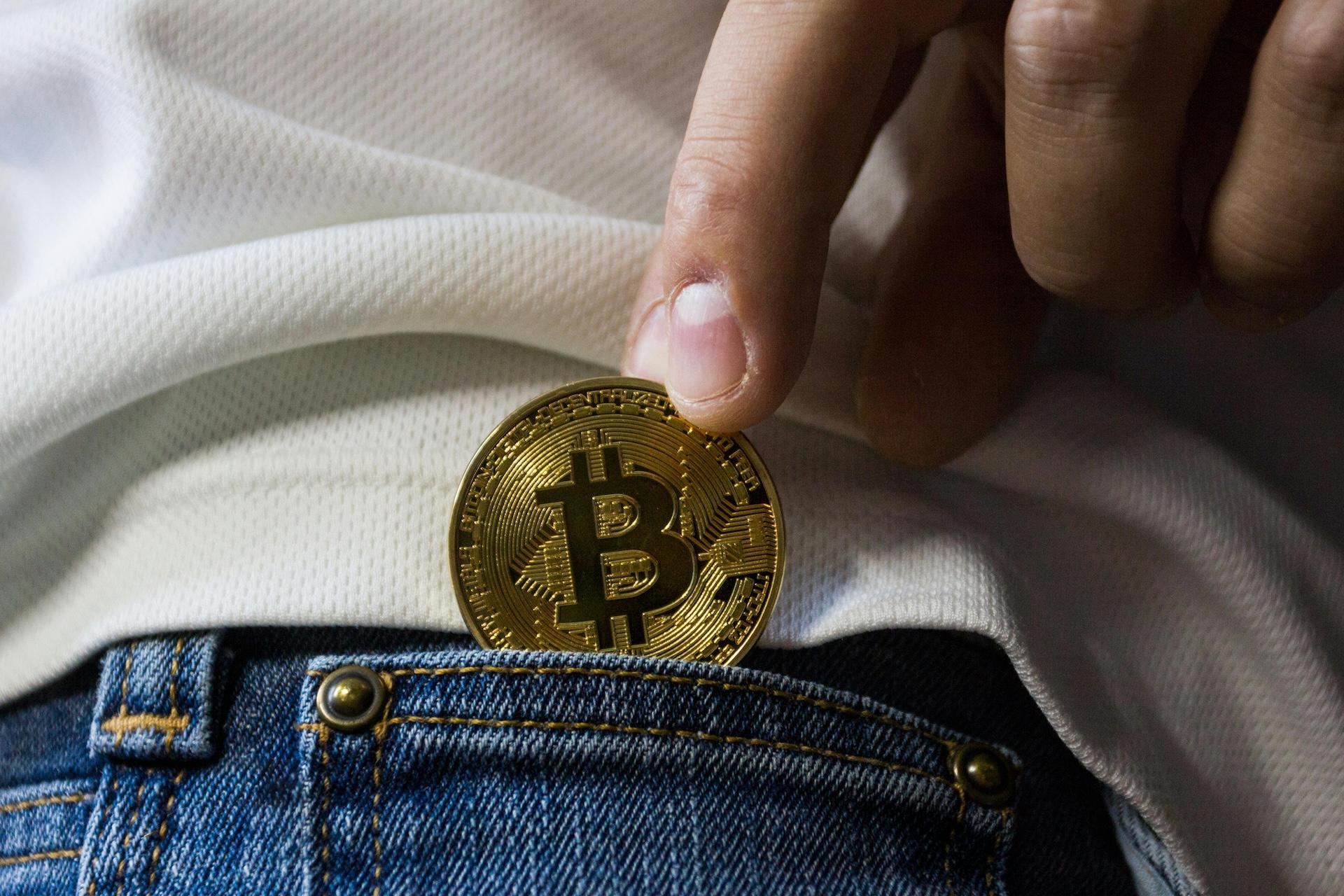 A close-up shot of a hand holding a gold-colored Bitcoin coin over a denim pocket. The background shows white textured fabric, and the coin displays the distinctive Bitcoin "₿" logo with its characteristic circuit-board-like pattern etched into the metallic surface.