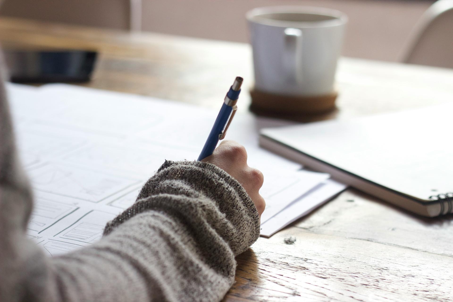 Hand in gray sweater sleeve writing on paper with blue pen, coffee cup in background