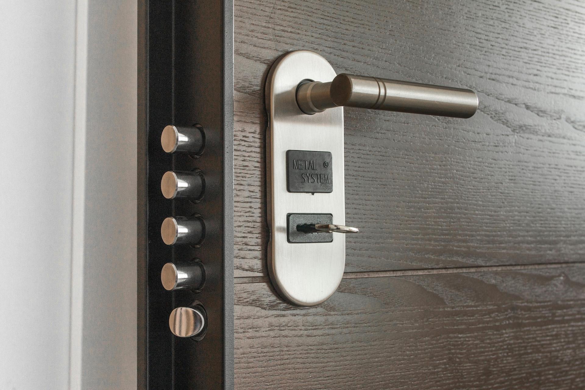 Modern door lock with metal handle and security system. The image shows door hardware consisting of a matte silver handle marked 'METAL SYSTEM', a keyhole with an inserted key, and a row of cylindrical metal bolts on a black plate on the left side of the door.