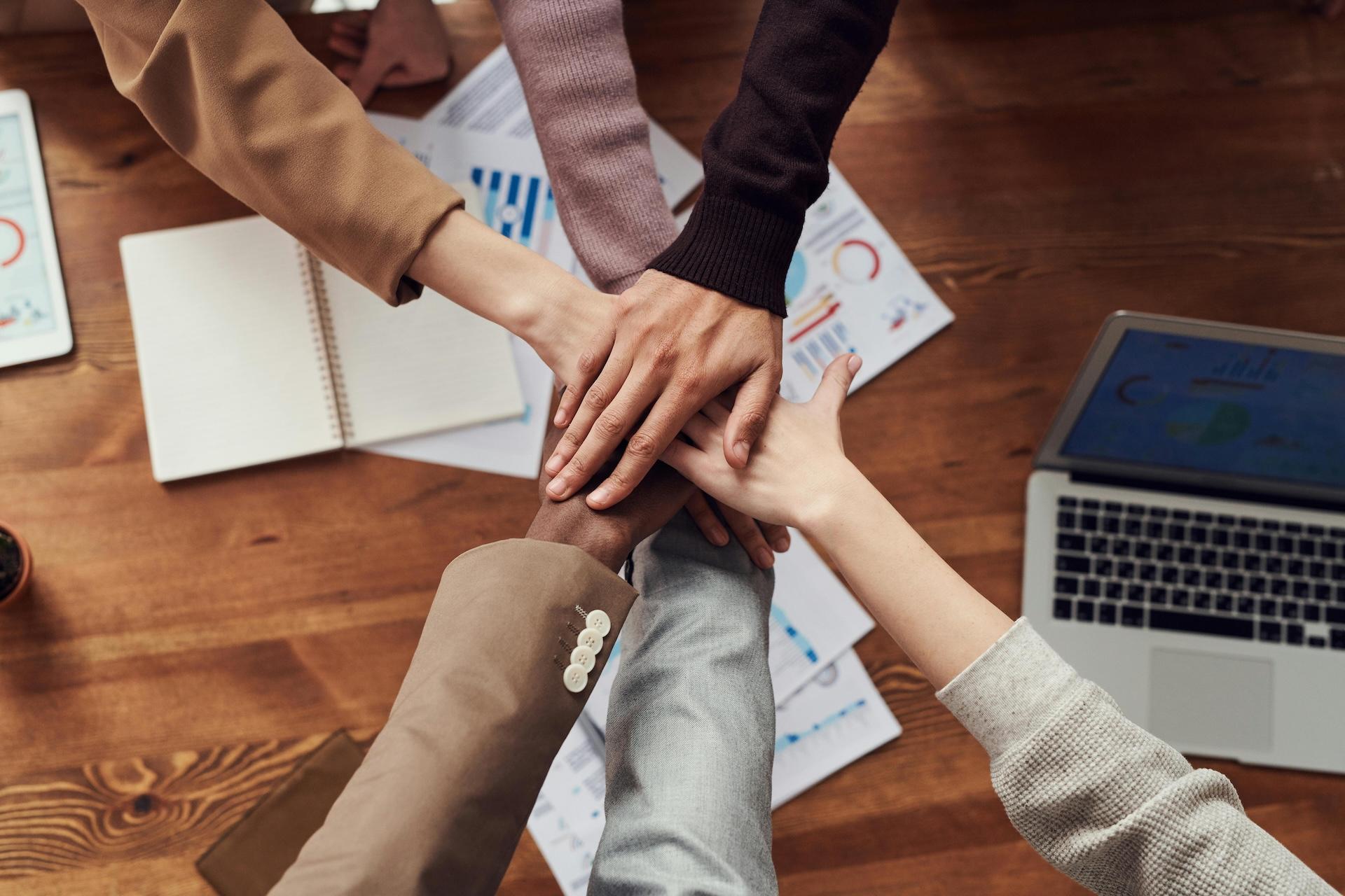 Overhead view of people putting their hands together in a teamwork gesture on a wooden desk. The desk surface has business documents with graphs, a laptop, and an open notebook.