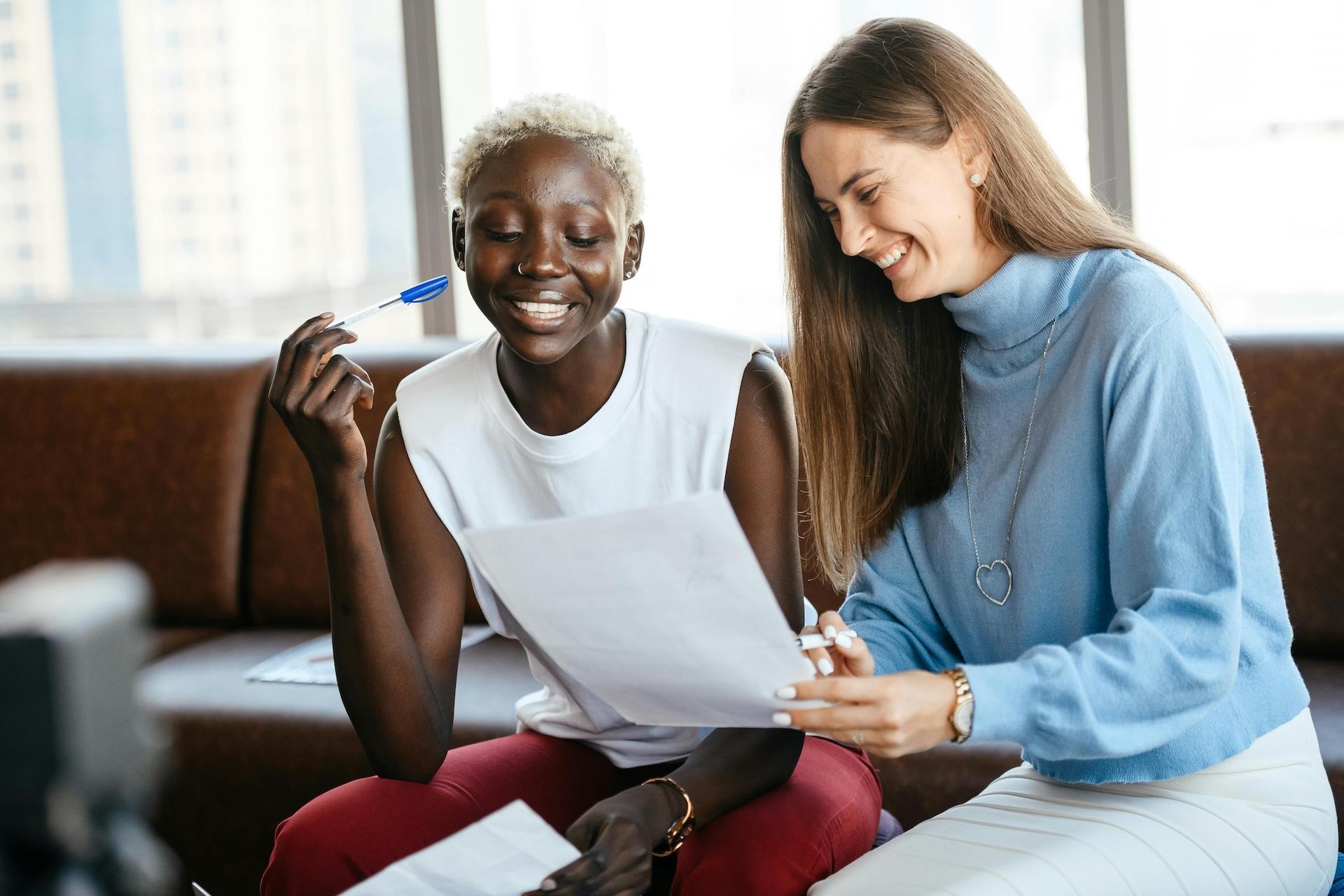 Two women sitting together and smiling while looking at documents, one wearing a white sleeveless top and burgundy pants, the other in a light blue turtleneck sweater.