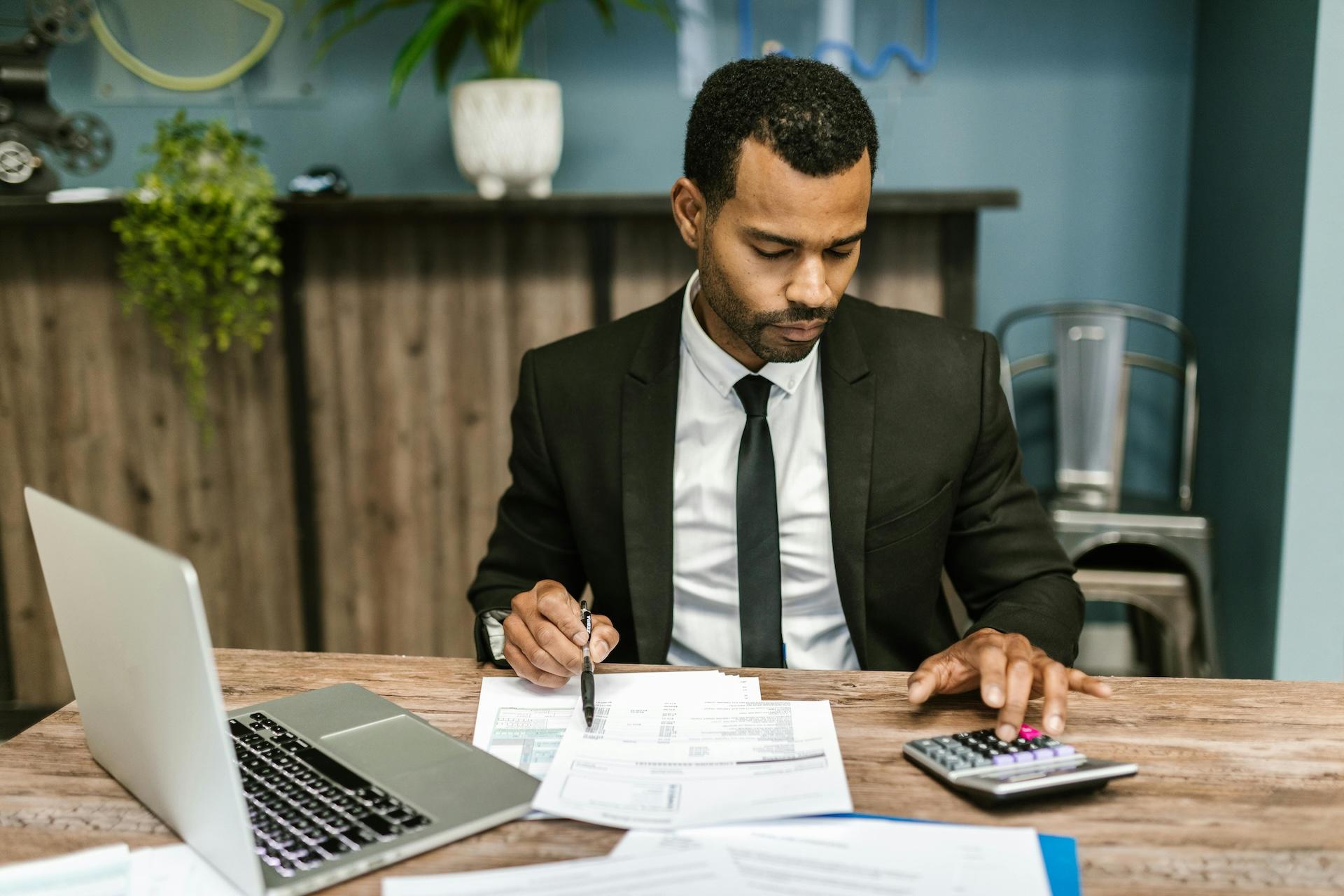 Businessman in suit working at wooden desk with laptop, documents, and calculator, looking focused while reviewing paperwork.