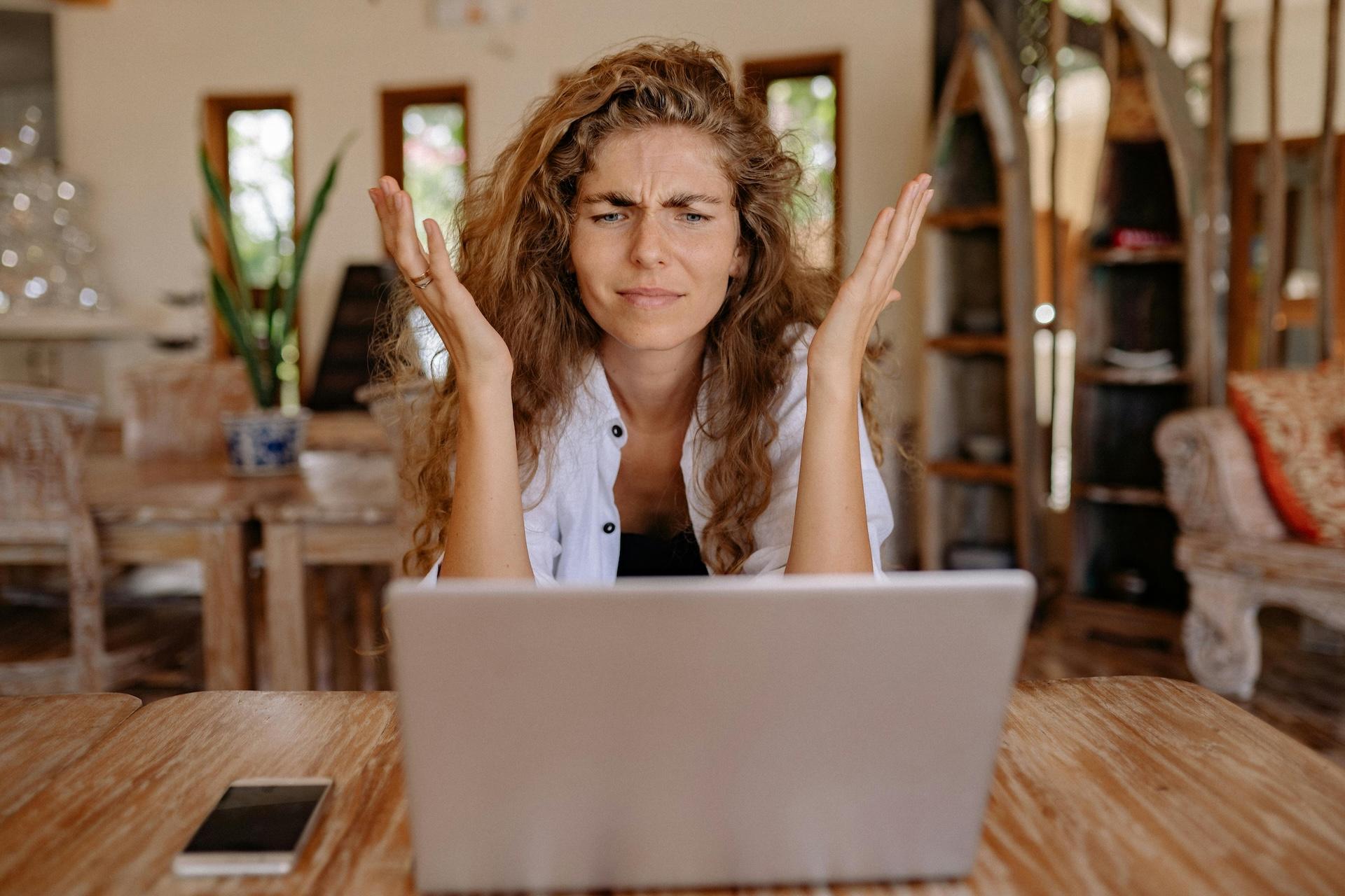 Frustrated woman with curly hair raising her hands in exasperation while sitting at wooden table with laptop. She's wearing a white button-up shirt and has a confused, annoyed expression on her face. The setting appears to be a home office or living room with house plants and wooden furniture in the background.