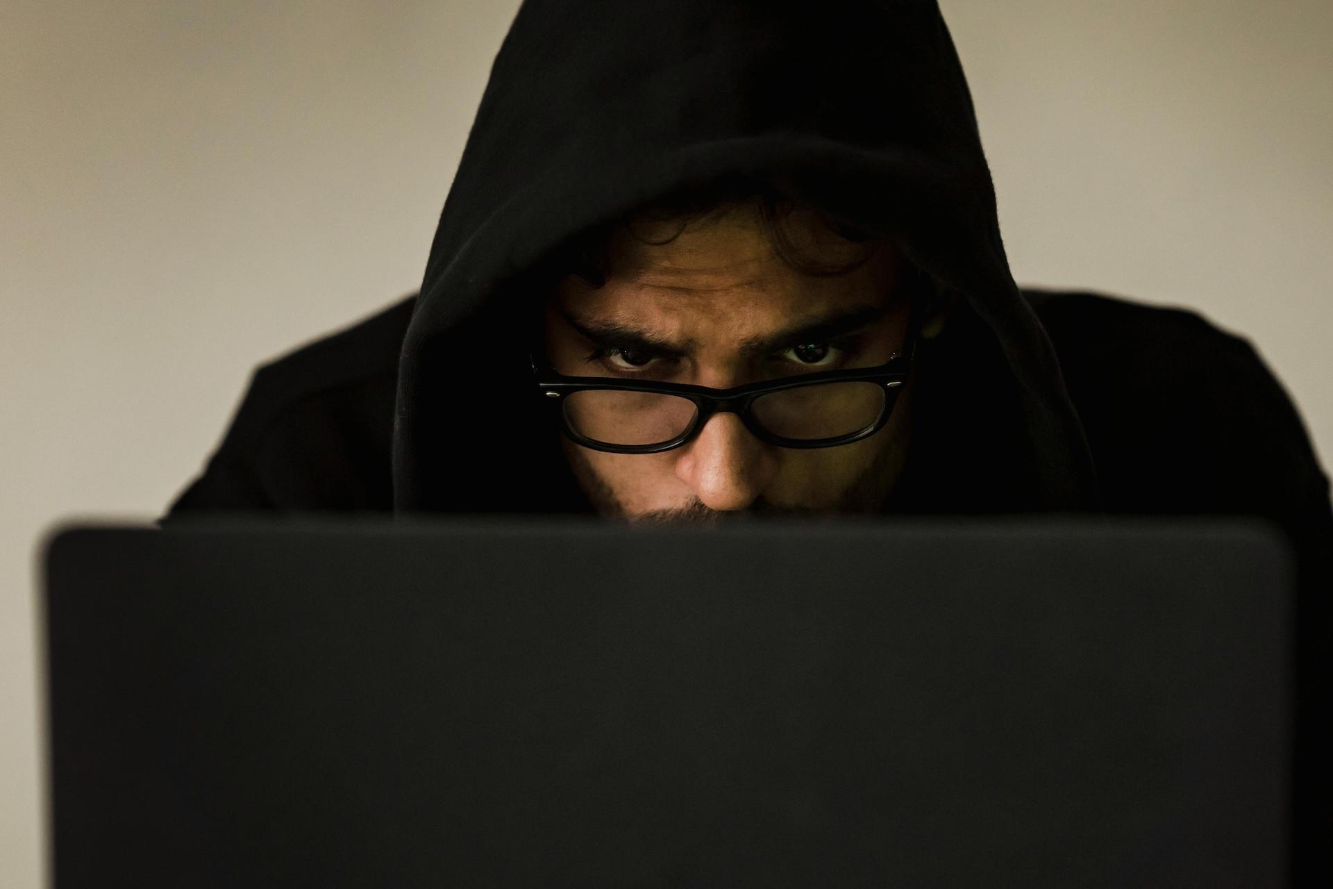 Person in black hoodie and glasses looking intently at laptop screen in dark room