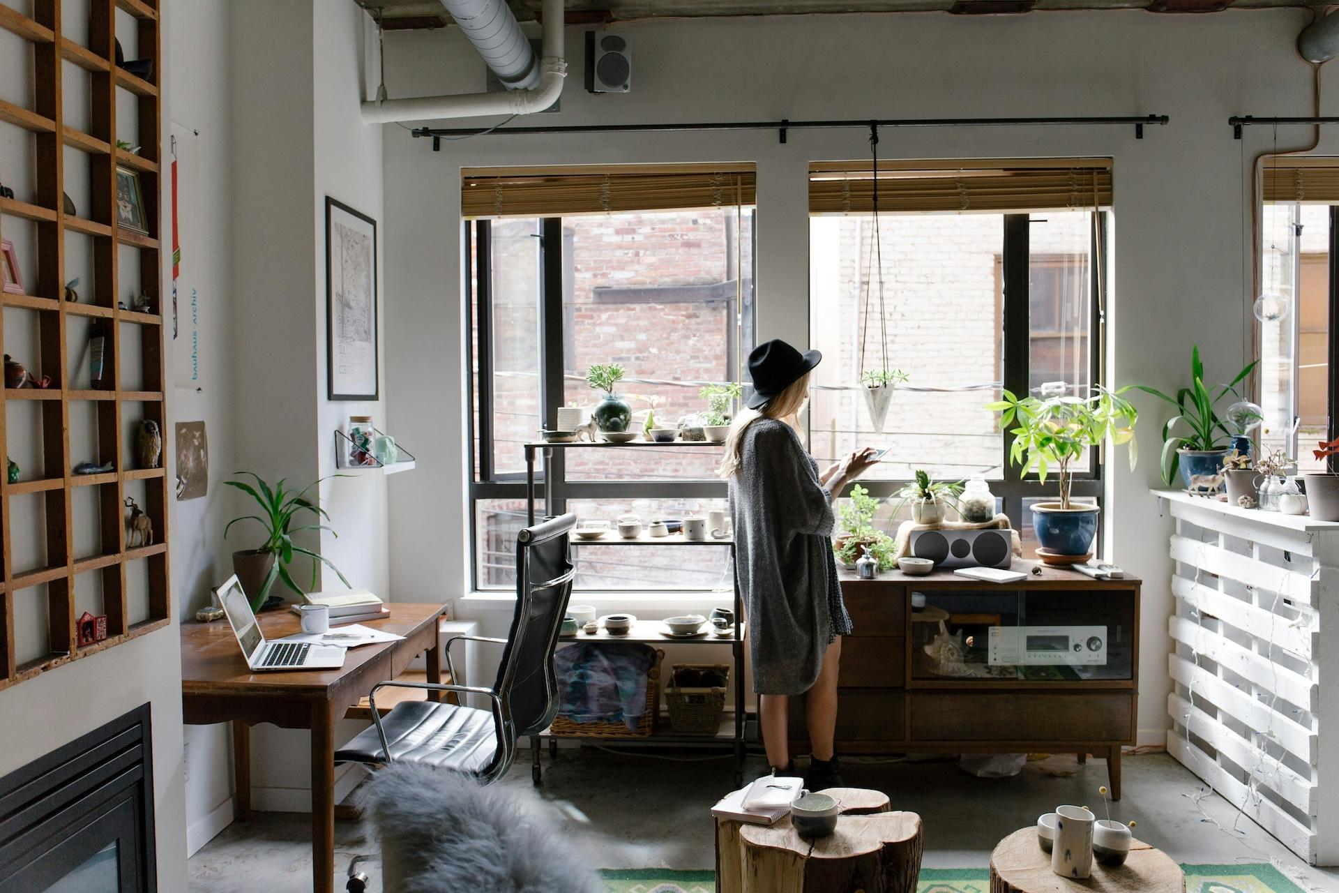 Cozy indoor space with a woman in a hat tending to plants near a window. The room features a desk with a laptop, wooden furniture, and various potted greenery.