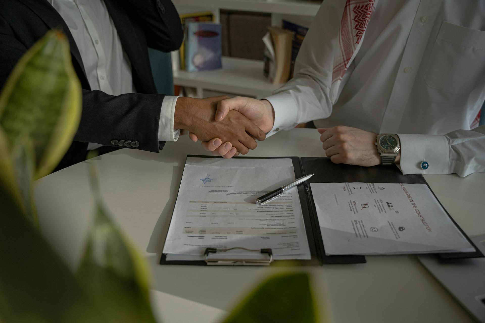 Two businesspeople shaking hands over paperwork on a desk, with documents and a pen visible in a professional office setting.