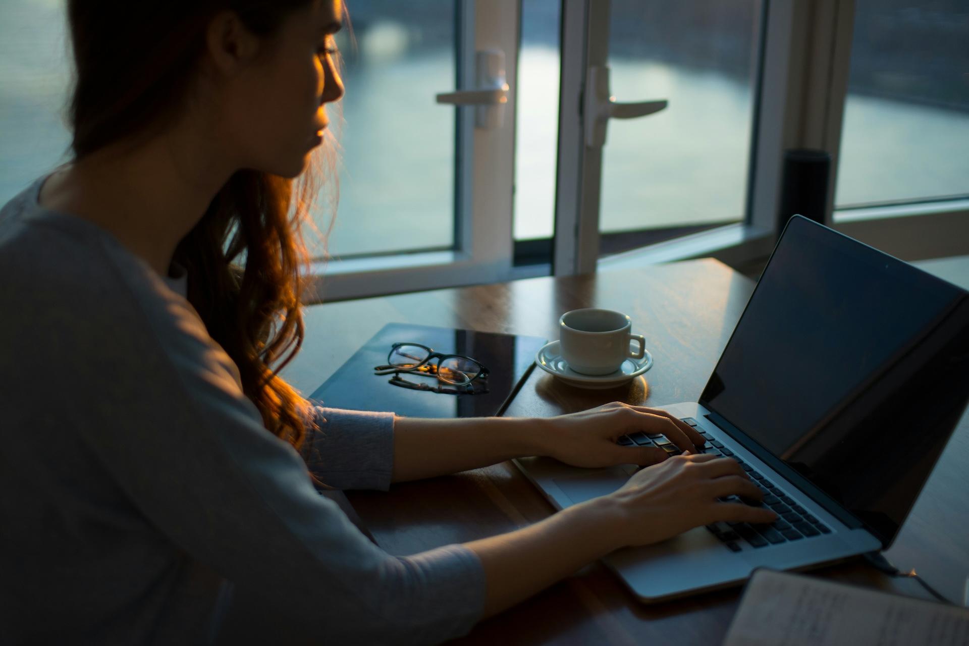 Woman typing on a laptop at a desk in low evening light, with a coffee cup and reading glasses nearby. The image is shot from the side, showing her silhouette against natural window lighting.